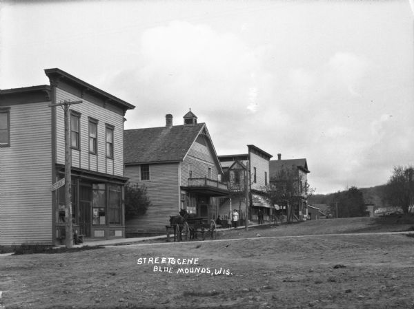 A view of a retail street from the intersection. From the corner of the street, there is a restaurant, hotel, and post office. On the sidewalk, men sit on benches, a woman and a child hold hands as they walk, and a man walks alone. A two-horse carriage stands at the curb.