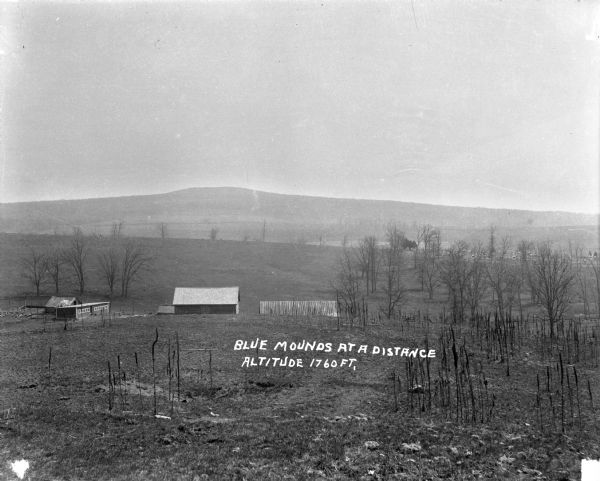 View looking down hill of several buildings. More hills are in the far background. Caption reads: "Blue Mounds at a Distance, Altitude 1760 FT."