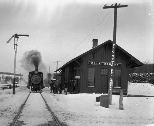 A steaming train at the depot platform on a snowy day. A group stands at the platform.