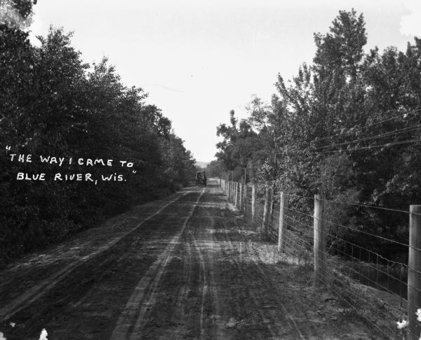 A dirt road with trees on one side and a fence on the other. A carriage is approaching.