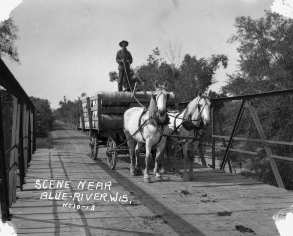 A man drives a team of horses pulling a wagon across a bridge. The man is standing atop the barreled goods, holding the reins. There is dung on the road.