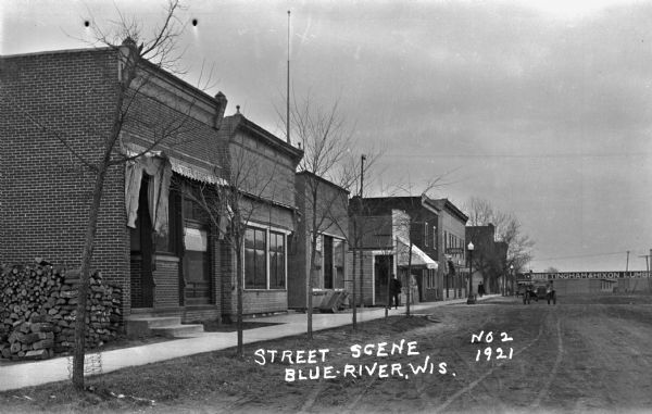 View from street of row of shops on along an unpaved street. A car drives on the street. A sign in the distances reads, "Brittingham and Hixon Lumber."