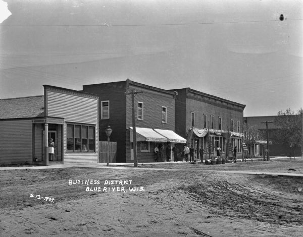 View of row of shops taken from the intersection. A public telephone station, post office, and a barbershop are on the street. Employees and pedestrians pose outside the shops. A dog sits on a chair outside the barbershop.