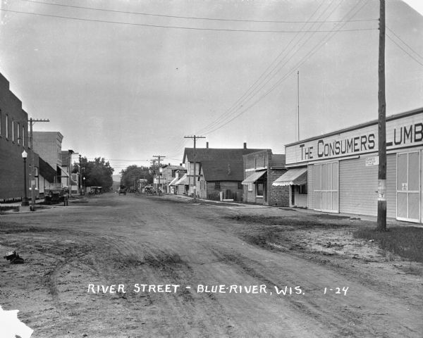 View down street. On the right is a sign for "The Consumers Lumber", among other small shops. A carriage is traveling along the street. A briefcase has been placed on the street, perhaps belonging to the photographer.
