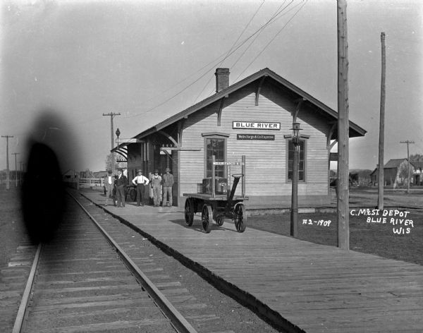 Exterior shot of the Blue River depot. Five men are standing outside the building. There is a Wells Fargo cart on the platform.