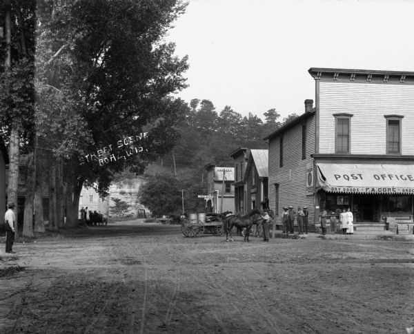 View down unpaved street with a grocery/post office at the corner. Horses are tied at the side of the store, near a group of men and women. A man stands on the far left.