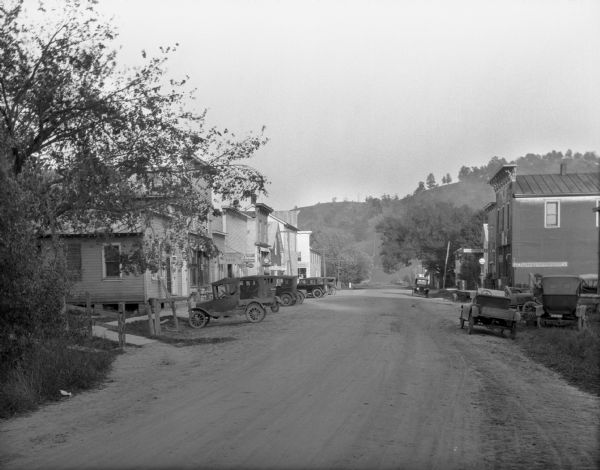 View down a dirt street. Cars are parked at the curbs along the shops. The shops include a post office, Ford repair shop, and furniture/rug store.