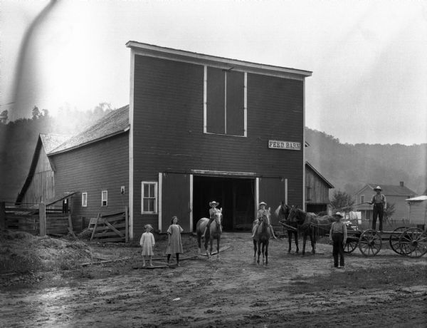 Two men, two boys on horses, and two girls pose in front of a feed barn.