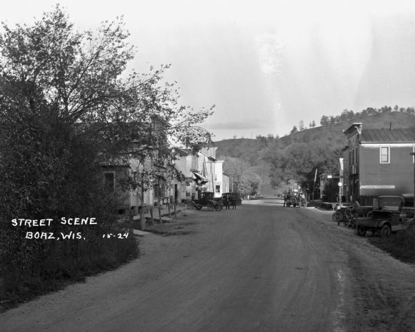 View down dirt street, with cars parked at the curbs near the shops. The shops include a post office, Ford repair shop, and furniture/rug store. Several men in suits are in the street.