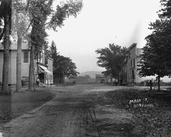 An intersection with a general store on the left. Two men stand outside the store. There are three horses and wagons on the right and one on the left.