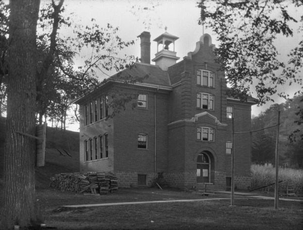 Exterior of the high school. The building features a bell tower, tall chimney, arched doorway and Dutch/Flemish gable. On the surrounding lawn, there is a log pile, seesaw (teeter-totter), and a still ring set.