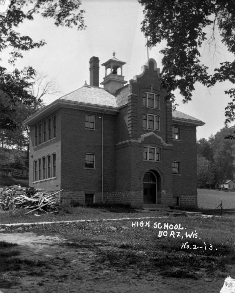 The exterior of the high school. The building features a bell tower, tall chimney, arched doorway and Dutch/Flemish gable. There is a hand-powered pump and stack of fuelwood on the property.