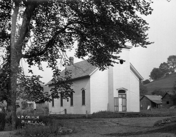 Exterior of church with tree in the foreground. One of the screen doors is fully opened. A dust pan hangs from a tree branch on the far left. There are homes behind the church.