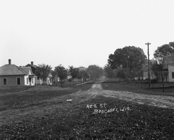 View down an unpaved residential road. Houses are in the background.