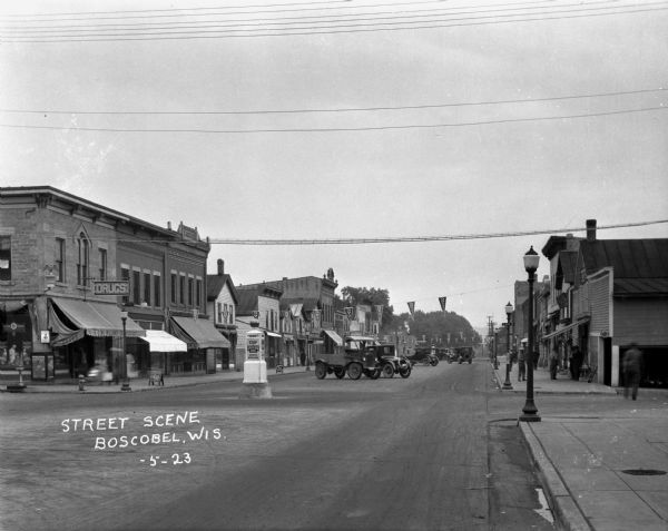 A retail street at the intersection of East Oat Avenue. Cars and trucks are parked in the middle of the street. The shops are: a drugstore, Guernsey's Bakery, a creamery, and Ford and Lincoln repair shop. Triangle flag-banners hang over the street.