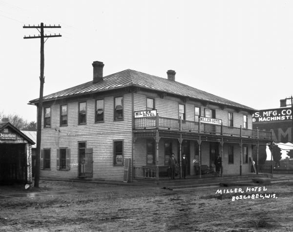 View from across street of three men standing on the porch of the Miller Hotel. There is an advertisement depicting a cow on the building next door.