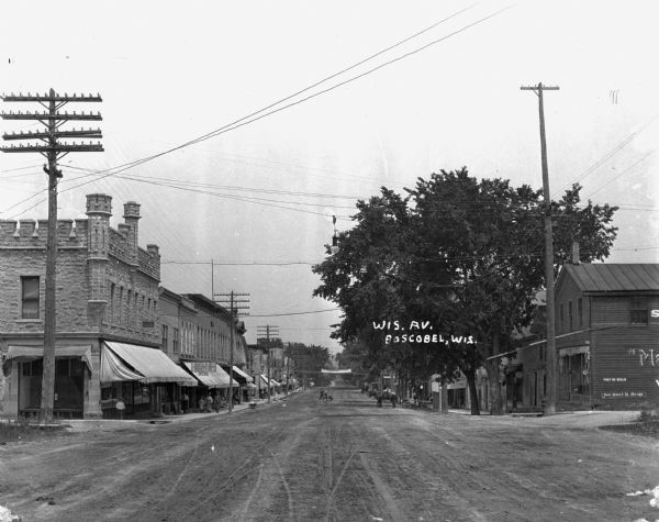 View down Wisconsin Avenue. Pedestrians are on the sidewalks, and there are shops signs for "Chas A. Blair - Lunch and Shortorders - News Stand." A banner across the street advertises the Boscobel fair.