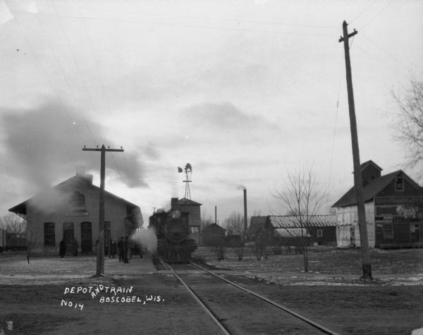 View from side of railroad tracks of a train at the Boscobel Depot on a snowy day. A crowd of people waits at the platform. There is a windmill in the distance.
