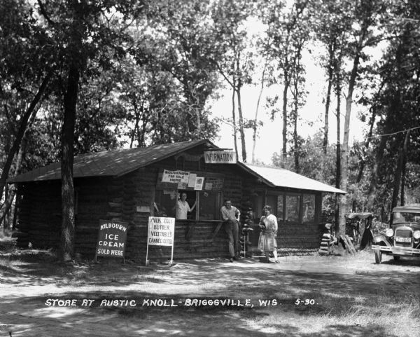 The log cabin store where a woman poses in the service window, and a man and a woman poses outdoors. There is a car parked in the road. The store advertises: Kilbourn Ice Cream, milk, eggs, butter, vegetables, canned goods, souvenirs, chicken dinners.