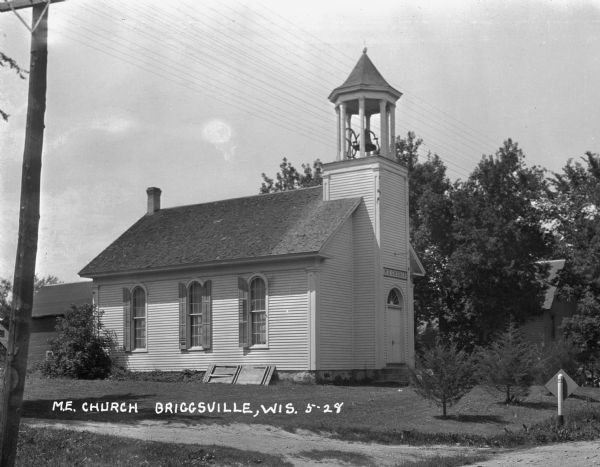 Exterior view of church building with a bell tower, arched windows, french doors, a fanlight, and a sign that read "M.E. Church."