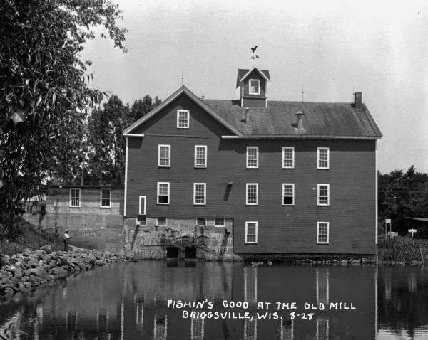A young man fishes on the rocky shore by the Old Mill. The Mill is three-stories high and features a cupola with a wind vane on top.