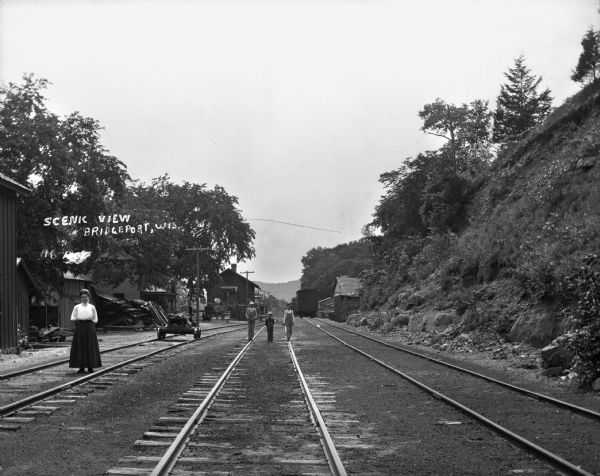 A view down railroad tracks near the Bridgeport depot. A woman with her hands behind her back poses on one track. Three boys wearing overalls pose on the middle track.