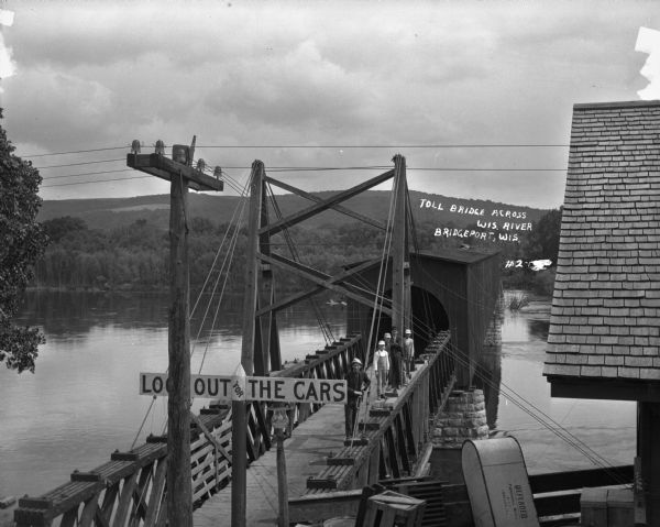Elevated view across the Wisconsin River. Men and boys stand on the railing of the covered bridge. A sign warns: "look out for the cars."