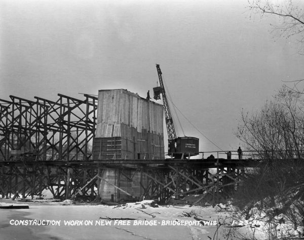 View from shoreline of a crane lifting machinery to workers at the top of the bridge during construction. Words on the crane reads, "Stevens Brothers and Shafer - Contractors - Saint Paul, Minneapolis."