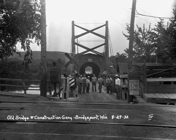Workers pose at the entrance of the old bridge. There are railroad tracks in the foreground with signs that read, "free bridge," "men working," and "caution - weight limit 4 tons - speed limit 10 miles."