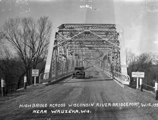 View down road of entrance of bridge. A car is driving towards the viewer. Signs at the bridge entrance read: "Parking on the Bridge is Prohibited," "Wisconsin River," "U.S. 18 35-61," and "You Are Entering Crawford County."