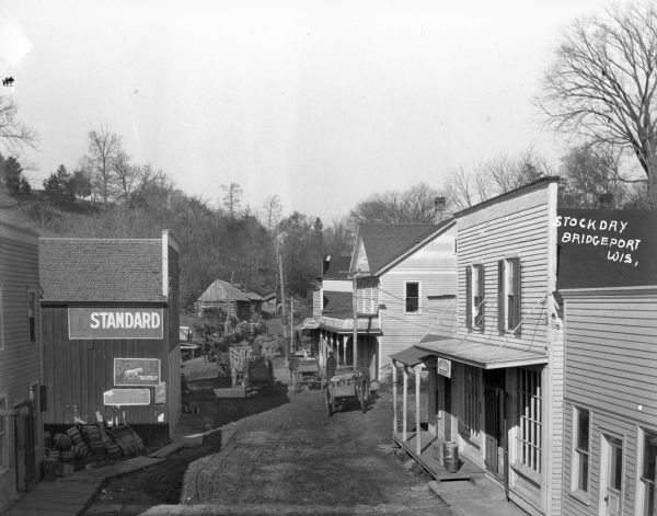Elevated view of the street on Stock Day. Horses and wagons are traveling down the street. The Bridgeport Hotel and the post office are on the right side. A general store on the left advertises Standard tobacco and Bickmore's Gall Cure.