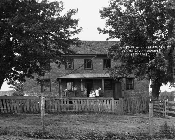View over fence of a family posing on the porch of a stone house. Text on photograph says: "Old Stone House Erected About 1800 for an Indian School."