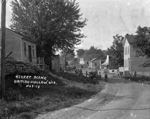 View down road of children posing on the side of the road. Two people sit in an automobile. In the background are commercial buildings, and horse-drawn vehicles.