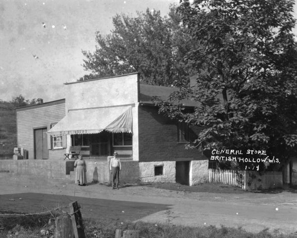 The exterior of P.J. Blindert's general store, with an awning over the storefront, and a loading dock on the left. A man and a woman stand in the road in front.