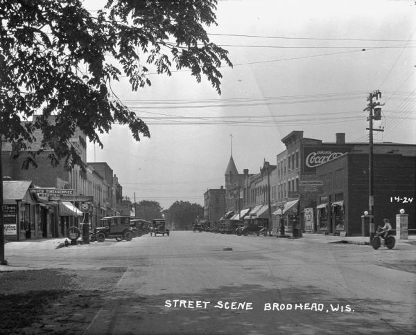 View down middle of street with many shops. Cars are parked at an angle along both sides of the street. A man rides a bicycle across the intersection. The stores include: Plumb and Timm Motor Company, United Tire and Service Station, Rexall drugstore, and a bakery/ice cream shop.