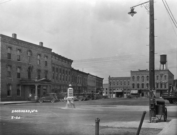 A street in the business section of Brodhead. Cars are parked along the curb near storefronts. In the foreground in the middle of the intersection is a street sign pedestal that has advertisements on it, as well as the directional signs SLOW" and "RIGHT." The shops include: Shorb Hotel, Brodhead Hardware Company, the Public Library, a Masonic temple, a theater, and a billiards hall. There is a water tower on top of the public library on the right.