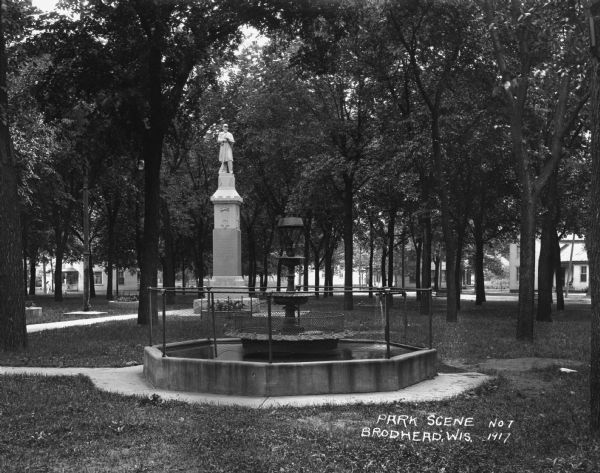View of a fountain in an octagonal basin at the Wisconsin Veteran's Memorial Park. There is a monument of a soldier behind it. Dwellings surround the park.