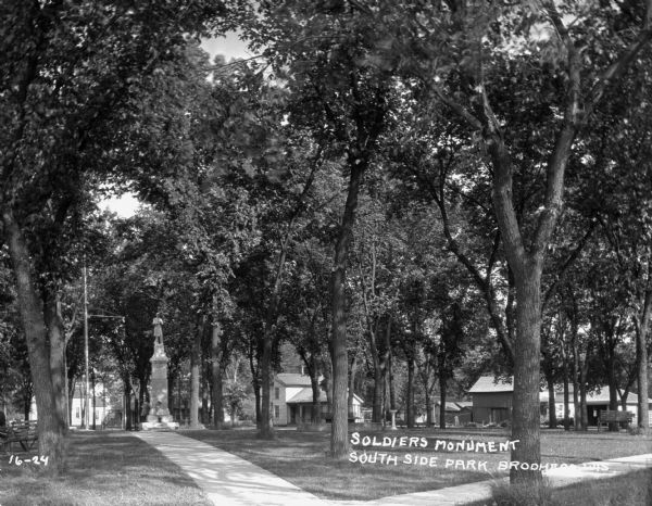 View down sidewalk of two young girls standing at the base of the Soldiers Monument. There is a fountain near the monument on the right. There are benches and trees in the park, and dwellings are along the street in the background.