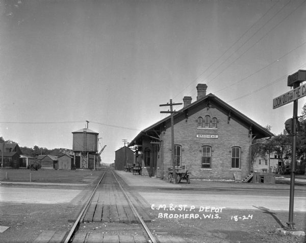 View down railroad tracks towards the train depot. There is a water tank on the left. Three carts are on the platform. A sign on the far right at the tracks says "Danger — Look out for the cars..."