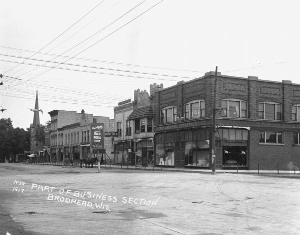 A retail street with the Terry Amerpohl Department Company, Will J. Smith — Rexall drugstore, a furniture store, and a church. A horse and buggy is parked at the curb by the drugstore. There is a Gold Medal Flour advertisement painted on the wall of a building.