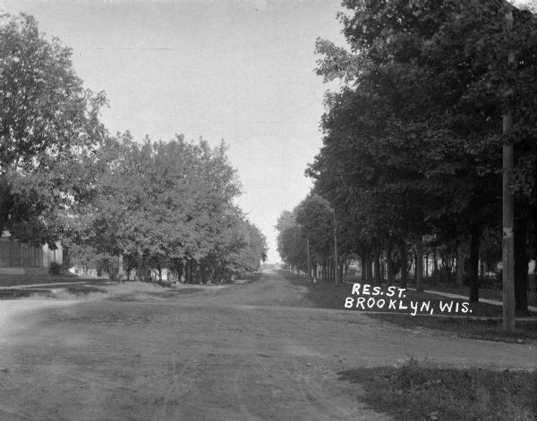 A view down a tree-lined dirt road in a residential area. Houses and sidewalks line both sides of the road, and pedestrians are in the far background.