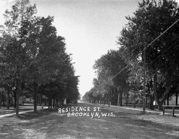 View down a tree-lined dirt road in a residential area. A car is parked at the curb in the distance.