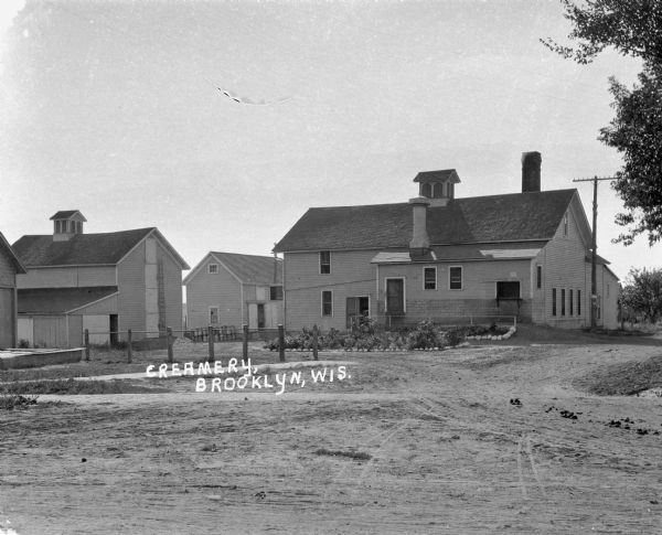 View from road of the exterior of the creamery. The main building features a gable roof, a cupola, two chimneys, as well as a garden in front. There are three more buildings, and stacks of milk cans are along the wall of one of them in the background.