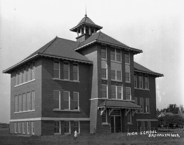 The high school building erected in 1908. The building features three stories, a bell tower, and a hip roof. A small child stands in front of the building.