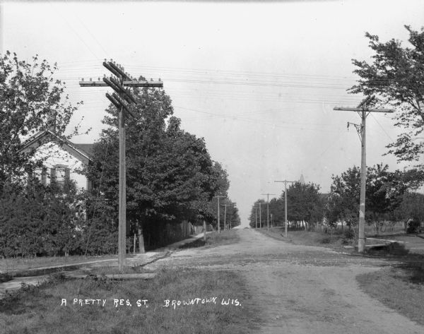 A view down an unpaved road lined with utility poles. On the left is a sidewalk and a house partially hidden by trees.