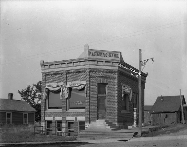 View from across street of the Farmer's Bank building on a corner. Behind the bank building are wood buildings, one of which has a sign that says, in part, "Krueg? Horse shoes."