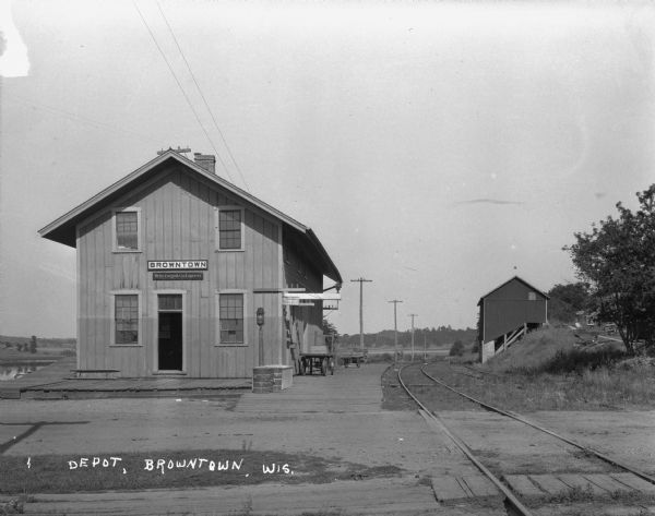 The Browntown train depot. The depot offers Wells Fargo & Company Express delivery and Western Union telegraph service. There are two carts on the platform. In the background on the right is a barn built on the side of a hill.