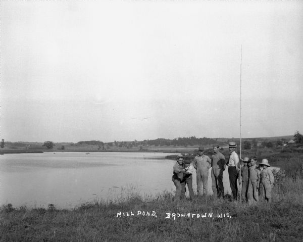 A group of men and boys pose by a pond. A boy holds a long, probably bamboo, fishing rod. Two men jokingly pose as if they are dancing the tango.