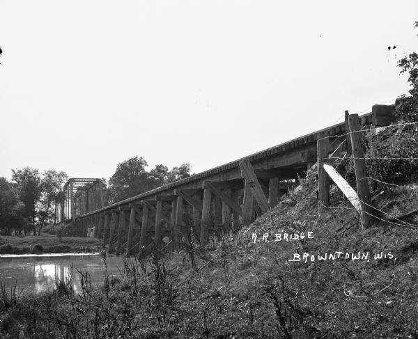 View from shoreline of railroad bridge across a river. Two girls and a boy are standing on the opposite side of the river near the shoreline.