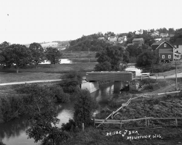 Elevated view of a stream with a dam and a bridge. There is a small fenced garden along the water. In the distance is a hillside with houses.